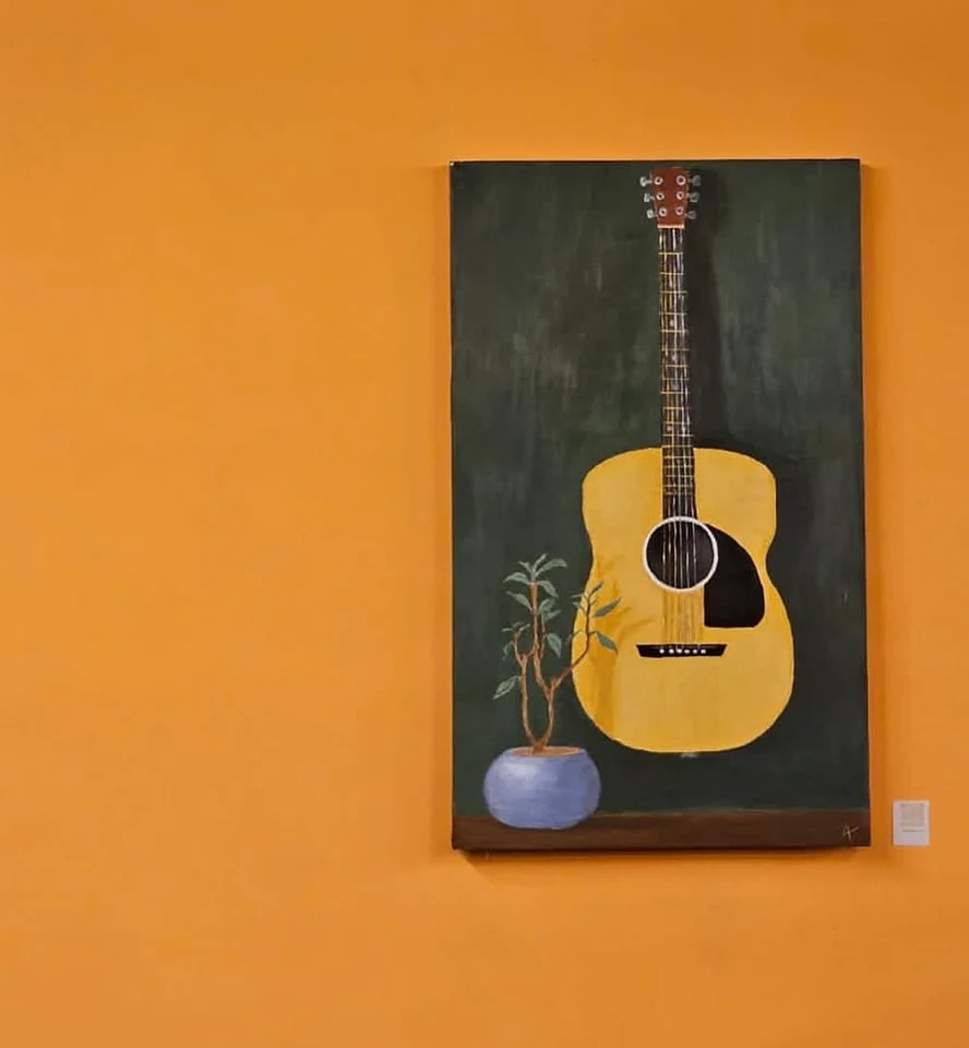 A yellow guitar and small plant on a dark panel displayed against an orange wall.